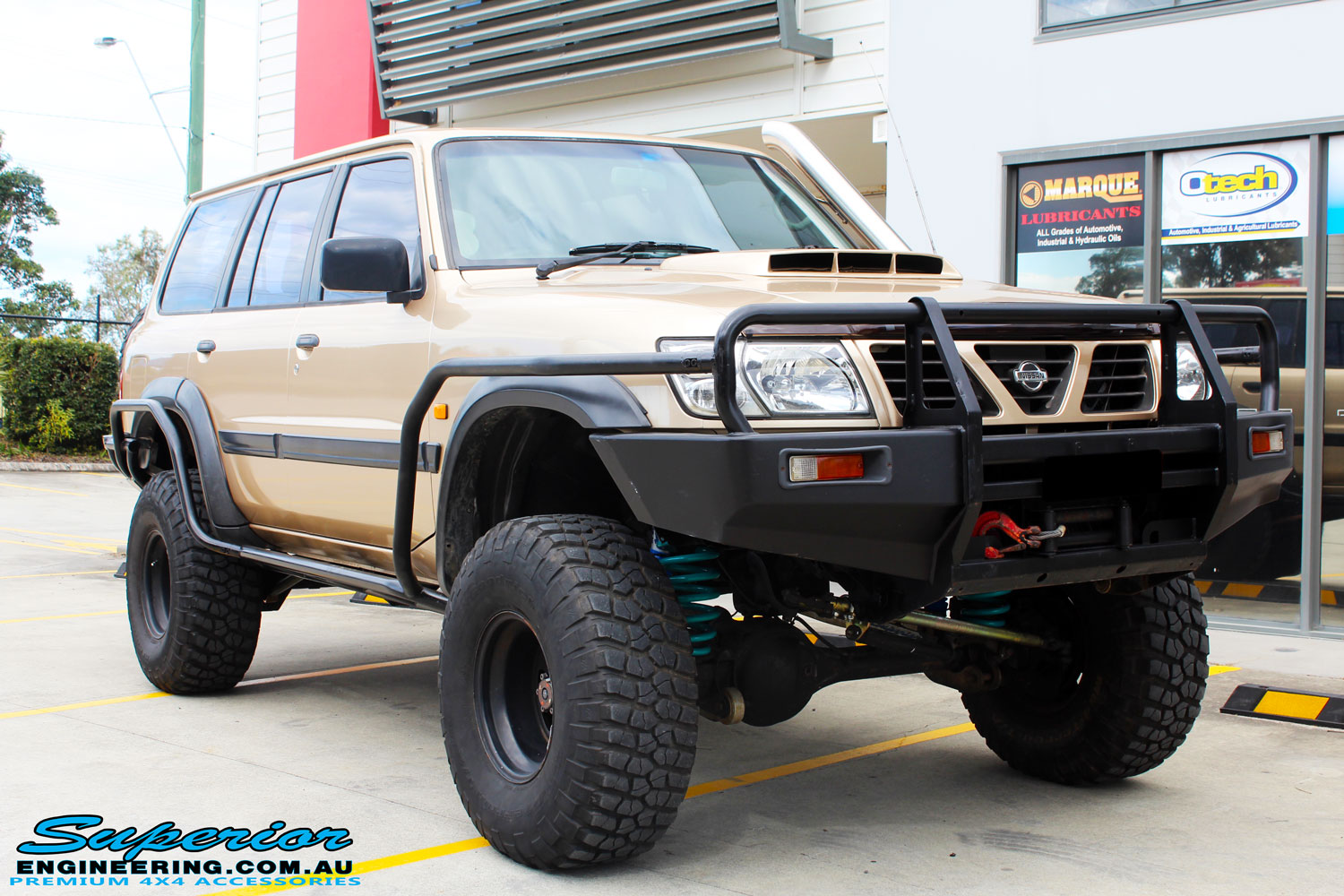 Right front side view of a Gold Nissan GU Patrol Wagon being fitted with Superior Lower Control Arms, Adjustable Upper Control Arms, Comp Spec 4340m Drag Link, 4" Inch Front & Rear Dobinson Coil Springs, Superior Coil Tower Brace and Superior 5" Inch Remote Reservoir Front & Rear Shocks